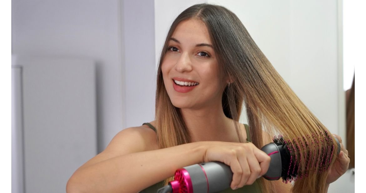 Jovem sorridente usando um secador de cabelo, ferramenta de calor, após aplicar um sérum com proteção térmica.